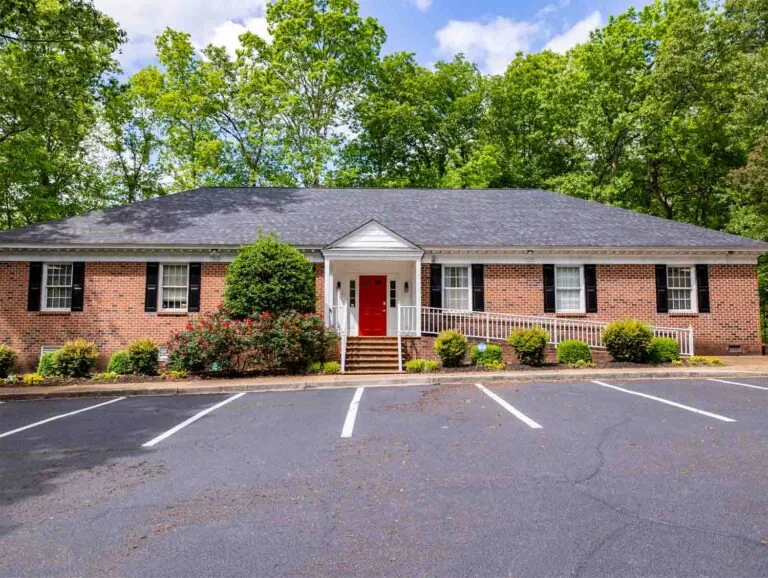 A brick building with a dark gray roof sits among trees and greenery, featuring a red front door, white trim, and black shutters. A parking lot with marked spaces is visible in front, and the scene is illuminated by natural light, creating a welcoming atmosphere.