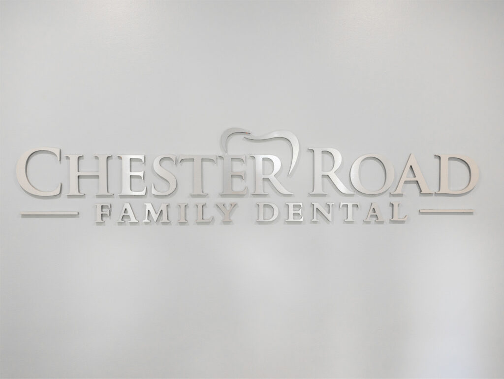 A metallic three-dimensional sign with the text "CHESTER ROAD FAMILY DENTAL." The design incorporates an outline of a tooth around the letters "R" and "O" in "ROAD."