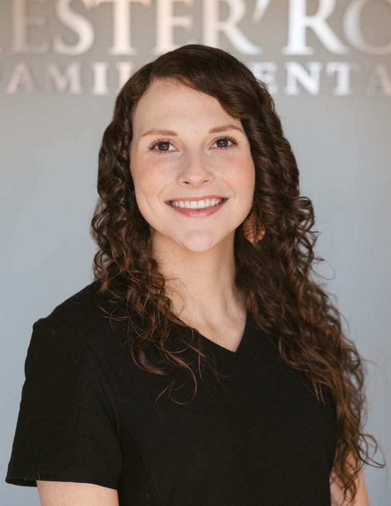 Headshot of Holly - Patient Coordinator with long, curly hair wears a black V-neck shirt with a background of Chester Road Family Dental.