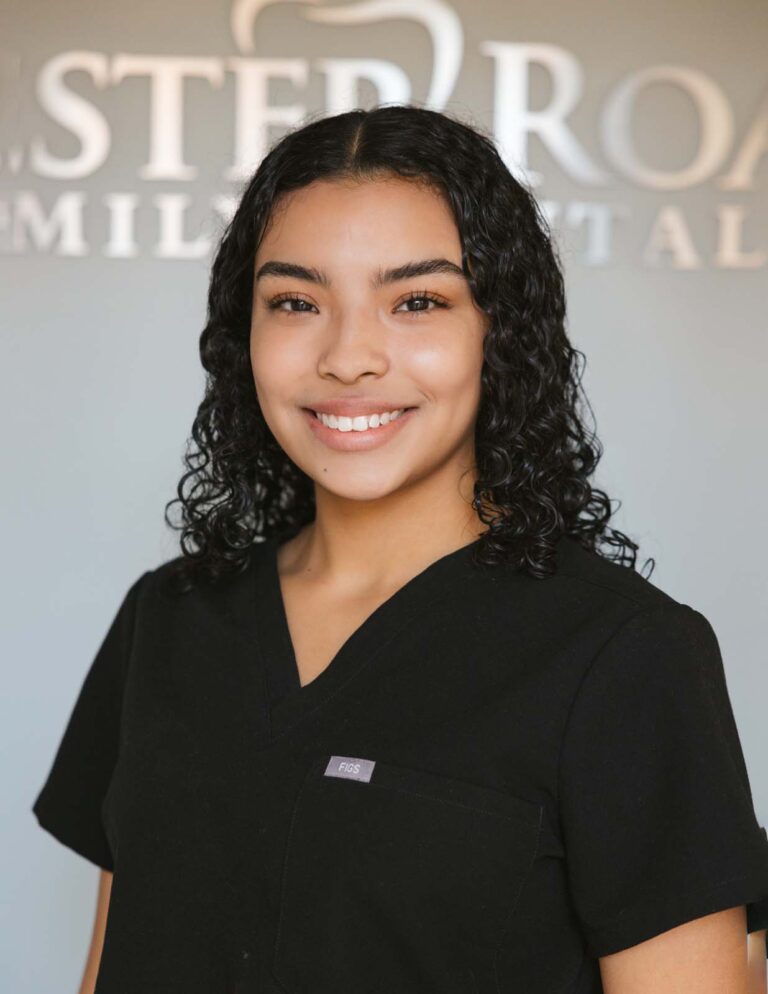 Headshot of Marilyn - Sterile Technician wearing a black scrub top with a visible logo, "Figs," on the chest pocket against a blurred background of Chester Road Family Dental logo.