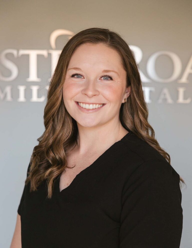 Headshot of Victoria - Dental Hygienist with long, wavy brown hair wears a black top against a blurred background of Chester Road Family Dental logo.